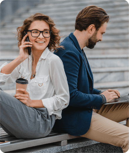 Two entrepreneurs sitting back to back outdoors, one on the phone with a coffee, the other on a laptop