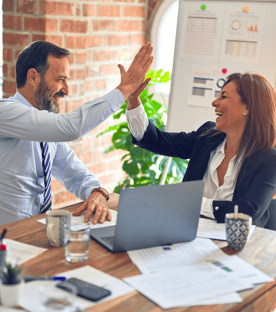 Two business partners high-five across a desk with a laptop