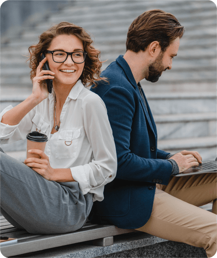 Deux entrepreneurs assis dos à dos en extérieur, l’une au téléphone avec un café, l’autre sur un ordinateur portable